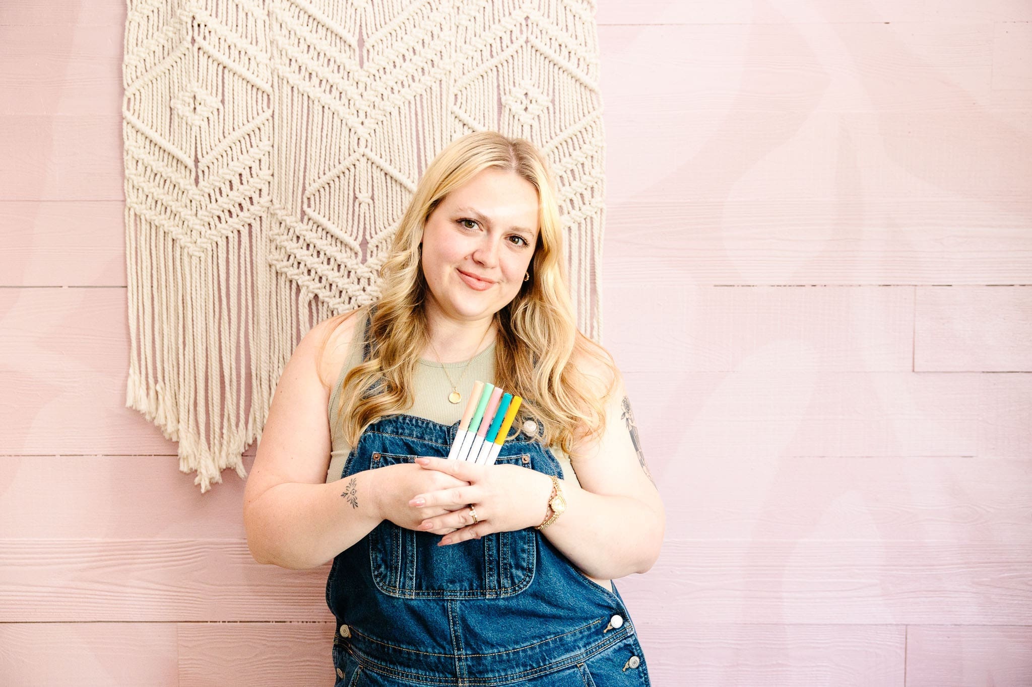Woman in front of pink wall and macrame project holds variety of colored markers and smirks at camera | Brand photos with Lafayette Lettering | Kayla Petrosky Photography