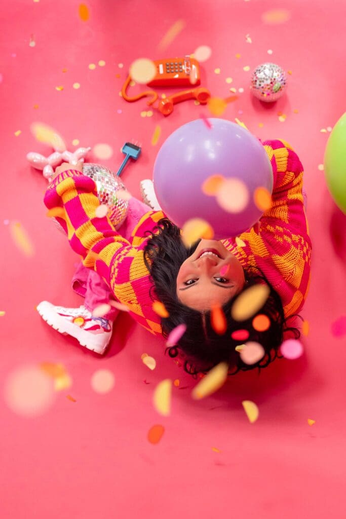 Woman holding balloons sits on pink backdrop and looks up as confetti falls around her | Balloon Artist Brand Photos
