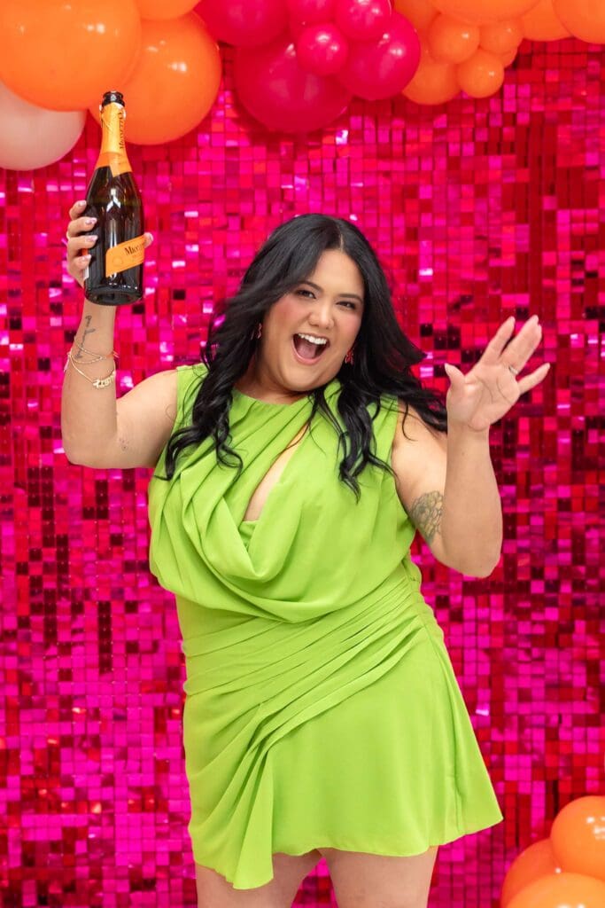 Woman in lime green dress dances in front of shiny pink backdrop while holding a bottle of bubbly | Balloon Artist Brand Photos