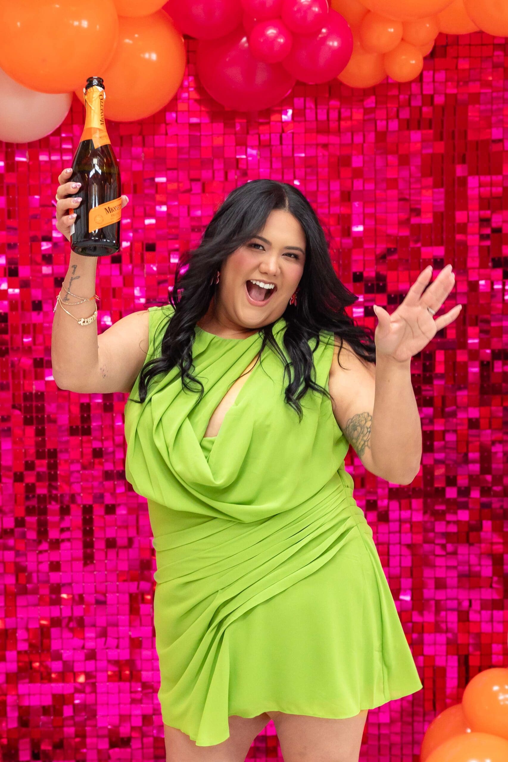 Woman in lime green dress dances in front of shiny pink backdrop while holding a bottle of bubbly | Balloon Artist Brand Photos