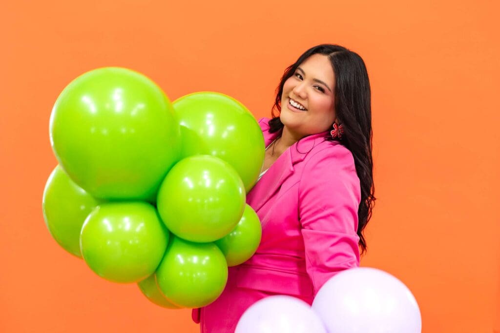 Woman in hot pink suit stands in front of orange backdrop while holding balloon arrangements | Balloon Artist Brand Photos